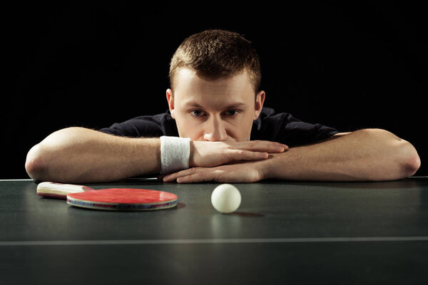 portrait of thoughtful tennis player at tennis table with ball and racket isolated on black