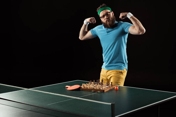 tennis player showing muscles while standing at tennis table with chess board isolated on black