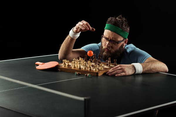 focused bearded tennis player throwing ball on chess board on tennis table isolated on black