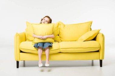 Little child with cushion in hands sitting on couch isolated on white clipart
