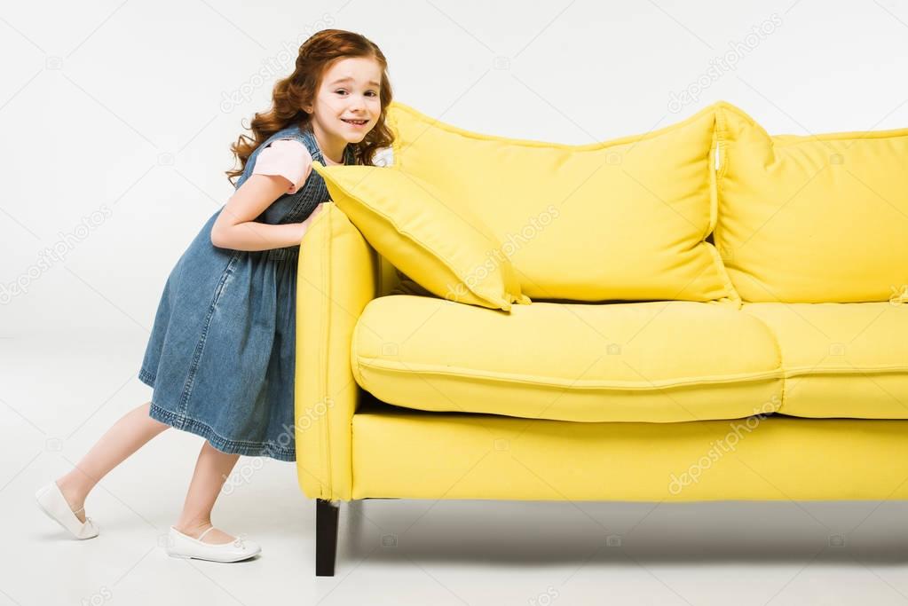Stylish little kid in dress pushing sofa isolated on white