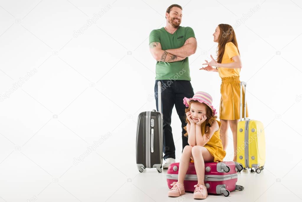 Upset little child sitting on suitcase with parents behind isolated on white, travel concept
