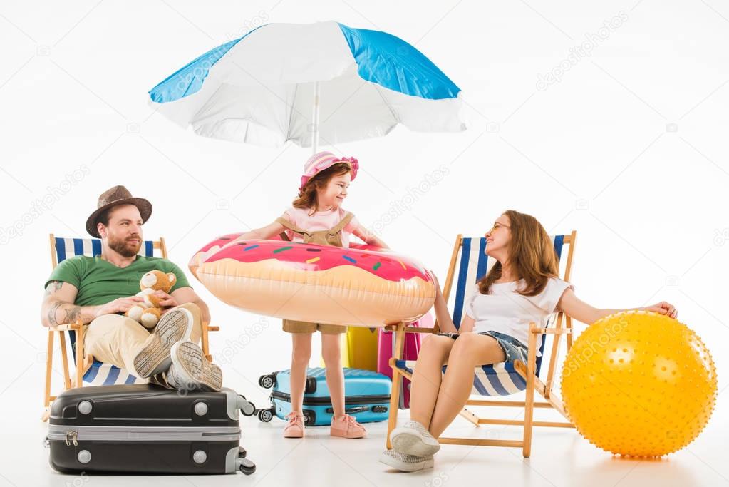 Happy little kid standing with flotation ring between parents sitting on sun loungers isolated on white, travel concept