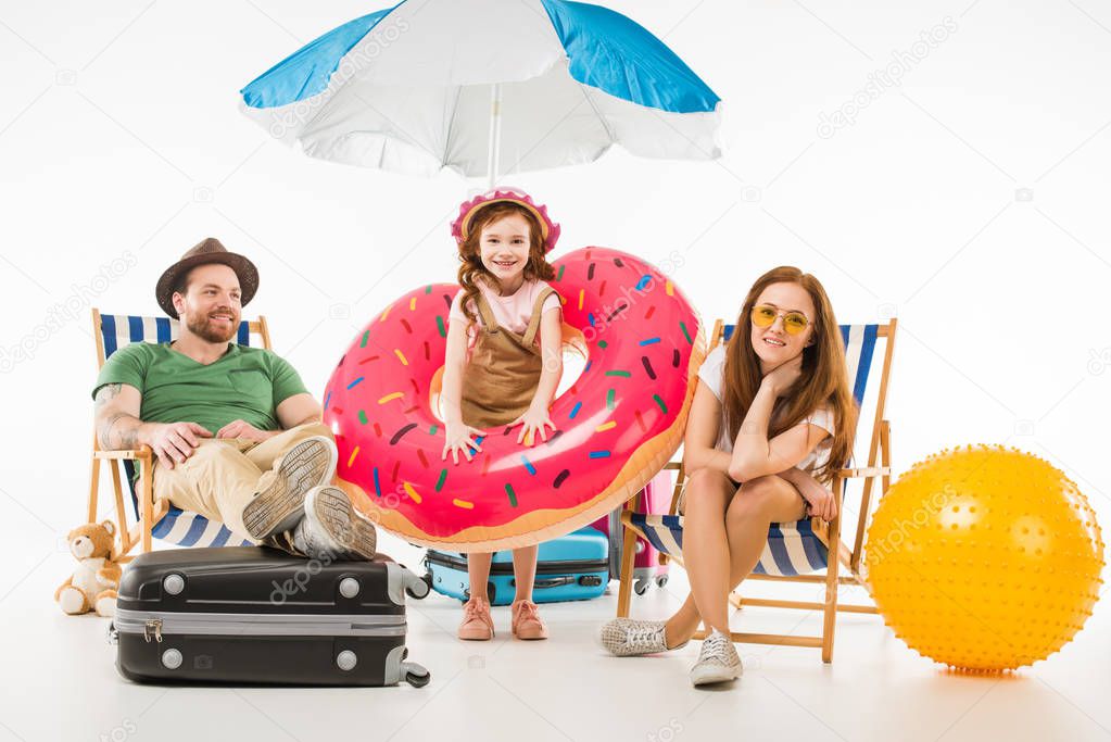 Little daughter with flotation ring standing between parents sitting on sun loungers isolated on white, travel concept
