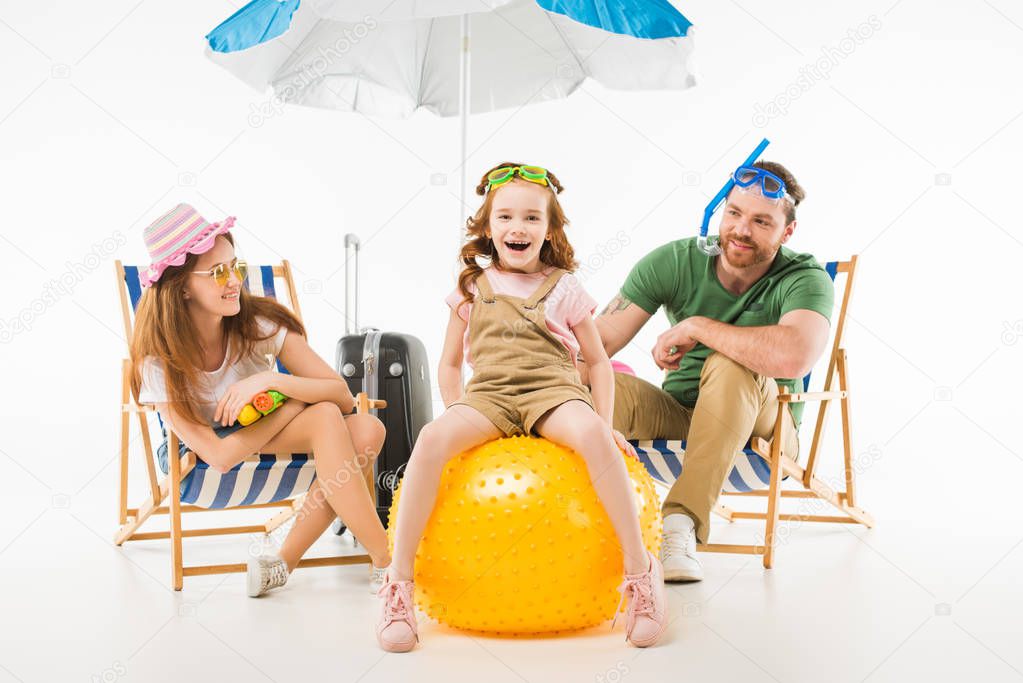 Happy little kid in swimming goggles sitting on ball between mother and father isolated on white, travel concept