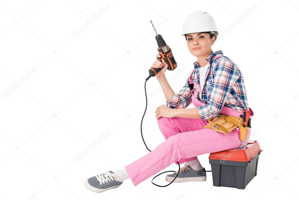 Professional builder in overalls and hardhat holding drill while sitting on toolbox isolated on white
