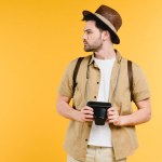 Young man in hat with backpack holding camera and looking away isolated on yellow