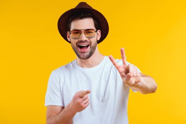 cheerful young man in white t-shirt, hat and sunglasses showing victory sign and pointing with finger isolated on yellow
