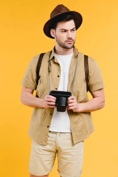 カメラと見て黄色の分離距離を保持しているバックパックと帽子でハンサムな若い男  — 無料ストックフォト