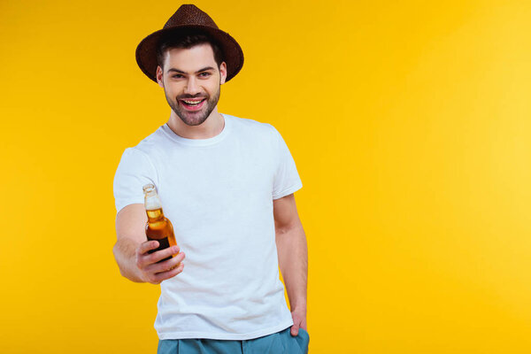 handsome young man in hat holding glass bottle of summer drink and smiling at camera isolated on yellow 