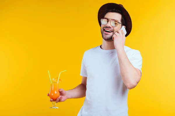 帽子とサングラス黄色の分離のスマート フォンでカクテルと話夏のガラスを保持でハンサムな笑顔の若い男 — ストック写真