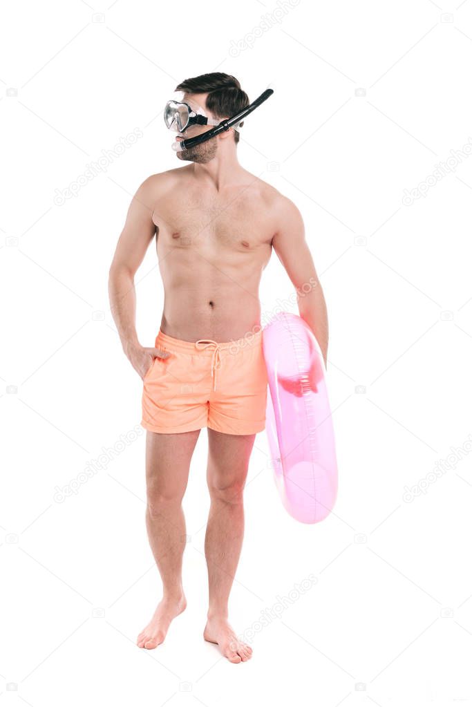 young shirtless and barefoot man in diving mask and shorts holding inflatable ring and looking away isolated on white