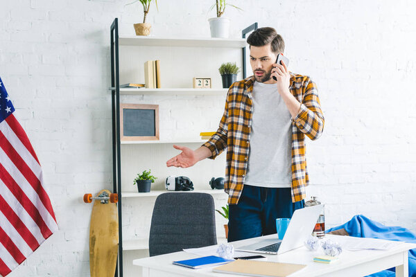 Freelancer standing by table and talking on phone in light office