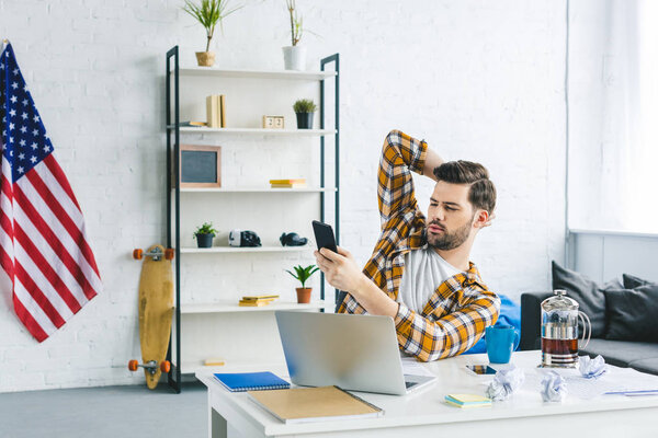 Freelancer working by laptop and holding smartphone in light office