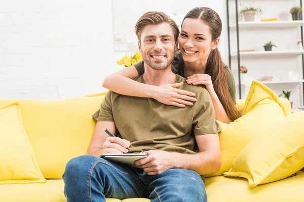 Joven Escribiendo Cuaderno Mientras Novia Abraza Por Detrás Sofá Casa — Foto de Stock