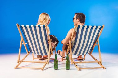 Smiling young couple relaxing in deck chairs by beer bottles on blue background clipart