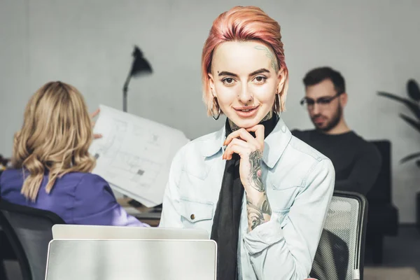 selective focus of smiling tattooed businesswoman at workplace with colleagues near by in office