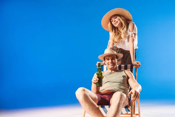 青の背景にビールを保持している麦わら帽子の男に椅子にもたれて金髪女性 — ストック写真