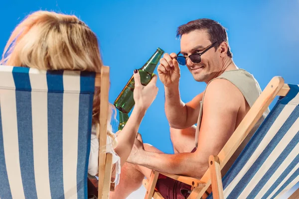 男子在太阳镜看着啤酒瓶在女性的手 而坐在沙滩椅上孤立的蓝色 — 免费的图库照片