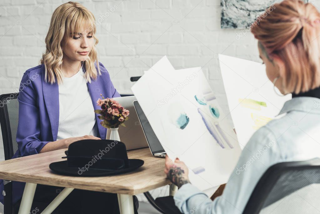 selective focus of tattooed designer looking at sketches at workplace with colleague near by in office