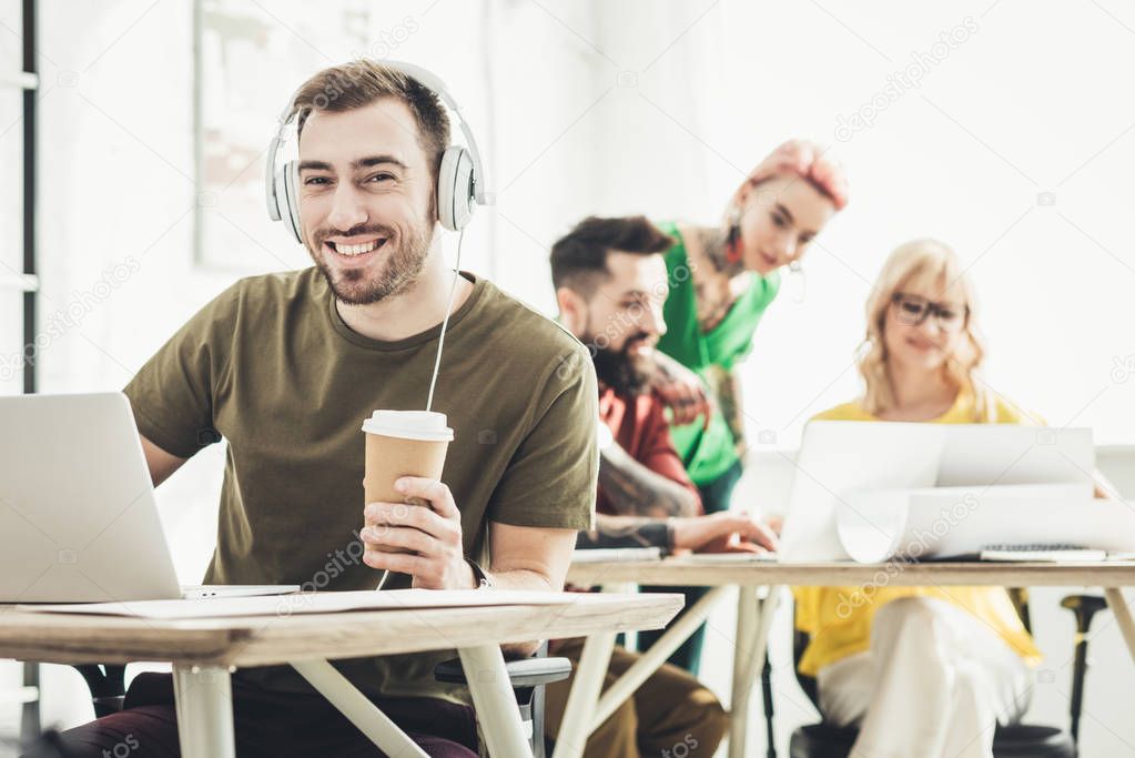 selective focus of smiling man in headphones with coffee to go and creative colleagues working behind in office