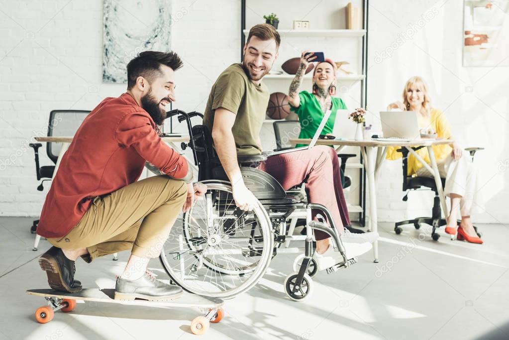 disabled businessman with laptop with colleague on skateboard and businesswomen behind in office 