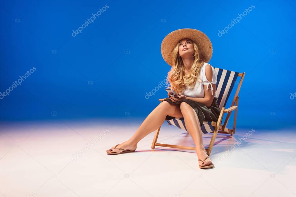 Blonde woman using smartphone and resting in deck chair on white background