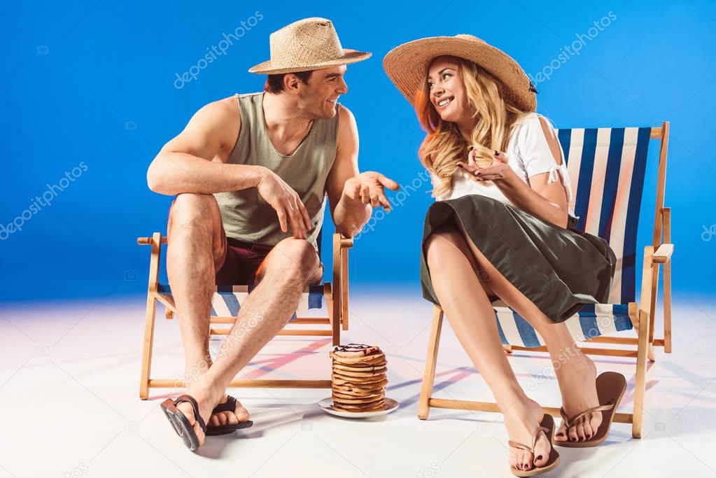 Smiling young couple talking and relaxing in deck chairs by stack of pancakes on blue background