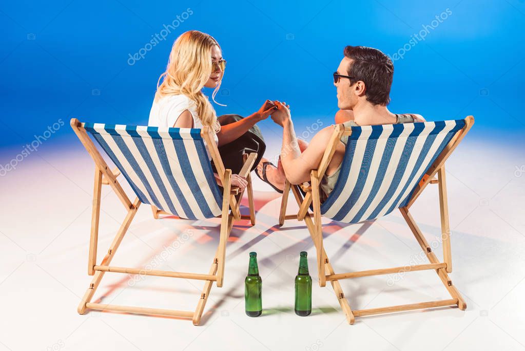 Young attractive couple holding hands in deck chairs by beer bottles on blue background