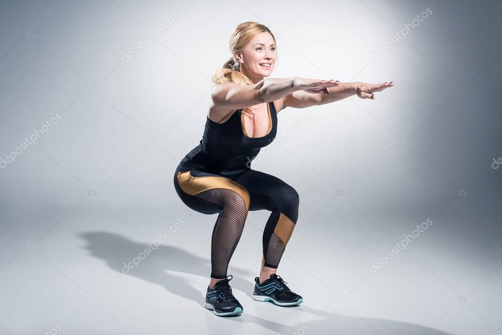 Smiling woman athlete performing squats on grey background