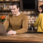 Sorridente giovane utilizzando il computer portatile mentre la ragazza cucina dietro in cucina