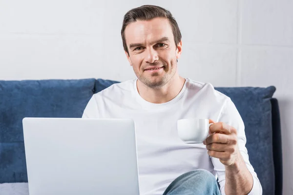Man Holding Cup Coffee Smiling Camera While Using Laptop Bed — Free Stock Photo