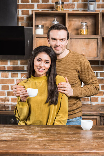 счастливая молодая многонациональная пара, обнимающая и улыбающаяся перед камерой, выпивая кофе на кухне
