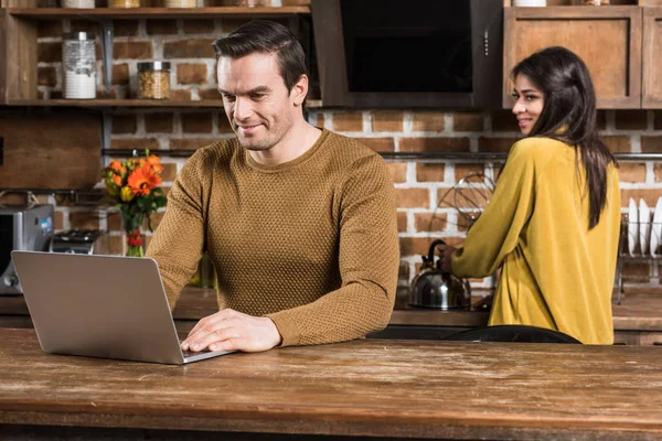 微笑的年轻人使用膝上型电脑 而女友在厨房后面做饭 — 免费的图库照片