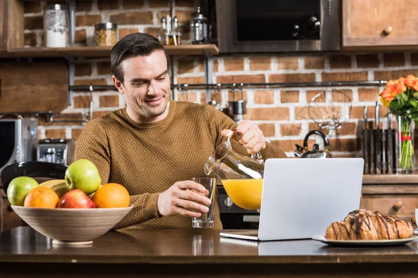 Bonito Sorrindo Homem Derramando Suco Enquanto Usando Laptop Mesa Cozinha — Fotos gratuitas