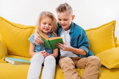 happy siblings reading book and sitting on yellow sofa