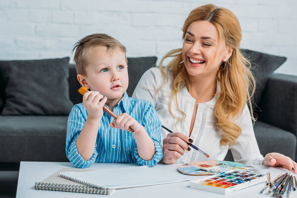 Happy mother and little boy with paintbrushes sitting at table