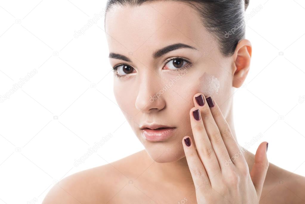 beautiful girl applying foundation cream on cheek and looking at camera isolated on white