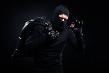 Man in balaclava holding plastic bag with cash and bitcoin clipart