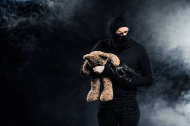Kidnapper in balaclava holding gun and teddy bear clipart