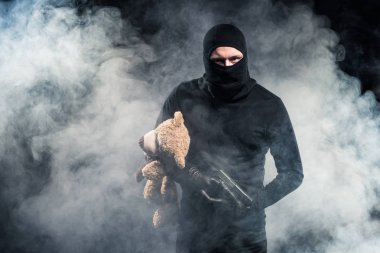 Criminal in balaclava holding gun and teddy bear in clouds of smoke clipart