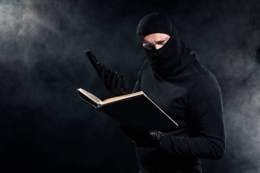 Man in black balaclava holding gun and reading book clipart