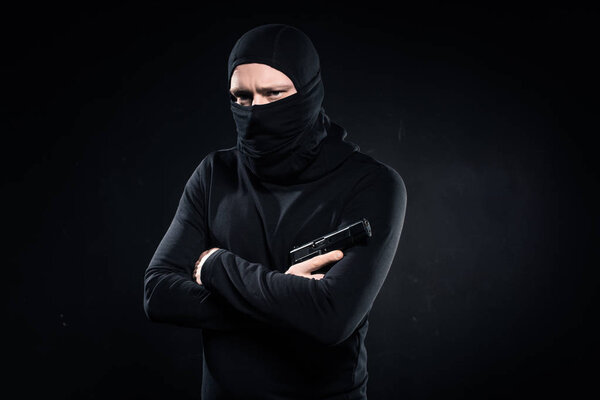 Thief in black balaclava holding gun in folded arms on black