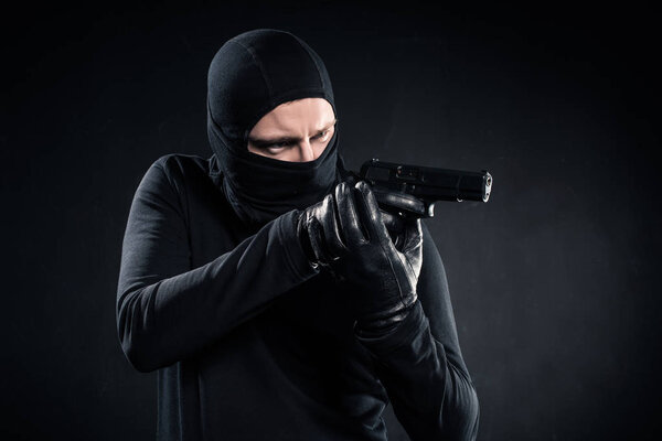 Burglar in balaclava aiming with gun on black