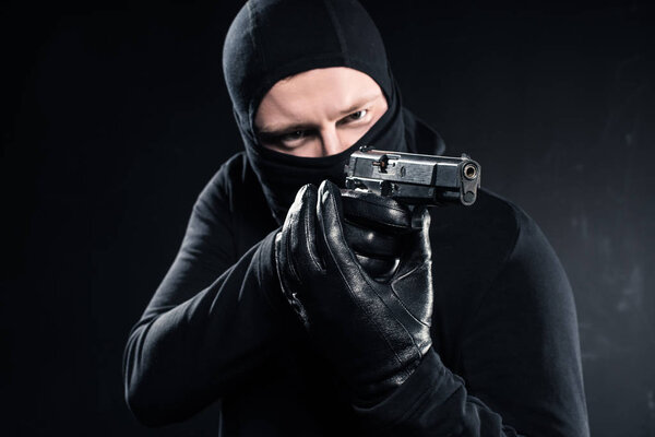Male robber in balaclava aiming with gun on black