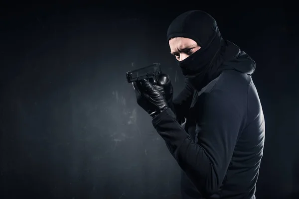Male criminal in balaclava and gloves aiming with gun on black