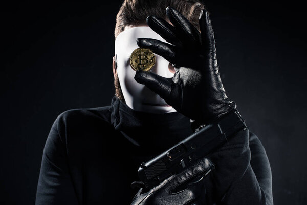 Thief in white mask holding gun and golden bitcoin