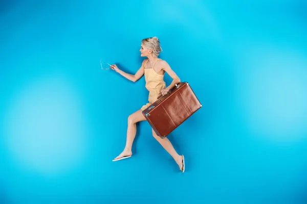 full length view of beautiful young woman in flip flops holding suitcase and running on blue