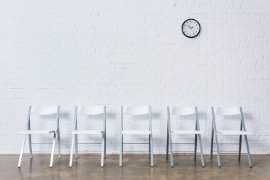Row of empty chairs by white brick wall with clock clipart