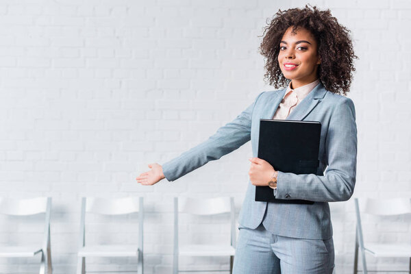 Young girl in suit holding folder and inviting to sit down on chair